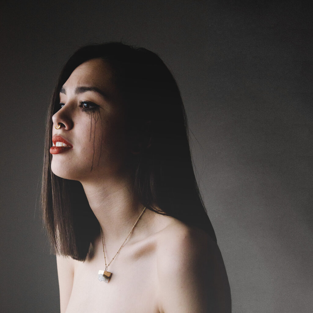 Andrea Gaspar a model with striking features poses against a muted background. Her makeup is bold with dark streaks running down her cheeks and her lips painted a vibrant shade of red. A delicate necklace hangs around her neck and she gazes upwards her expression contemplative.